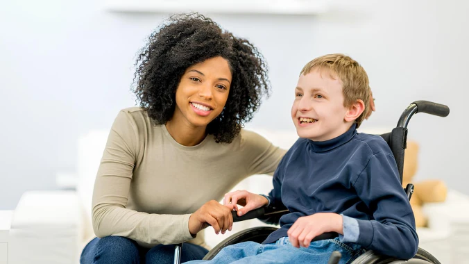 A smiling young woman with curly hair sitting next to a happy boy in a wheelchair, both enjoying a moment together in a bright, modern room.