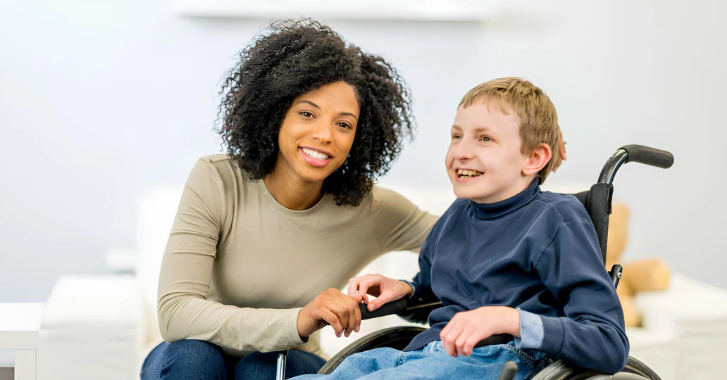 A smiling young woman with curly hair sitting next to a happy boy in a wheelchair, both enjoying a moment together in a bright, modern room.