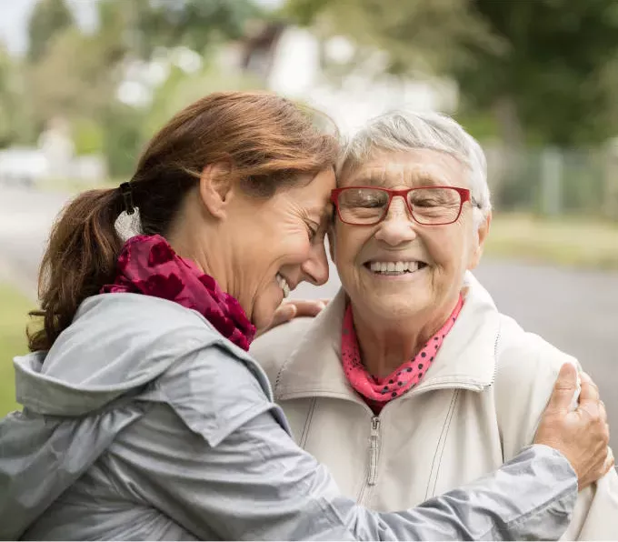 Smiling elderly woman with red glasses hugging a younger woman outdoors, both sharing a  joyful moment.