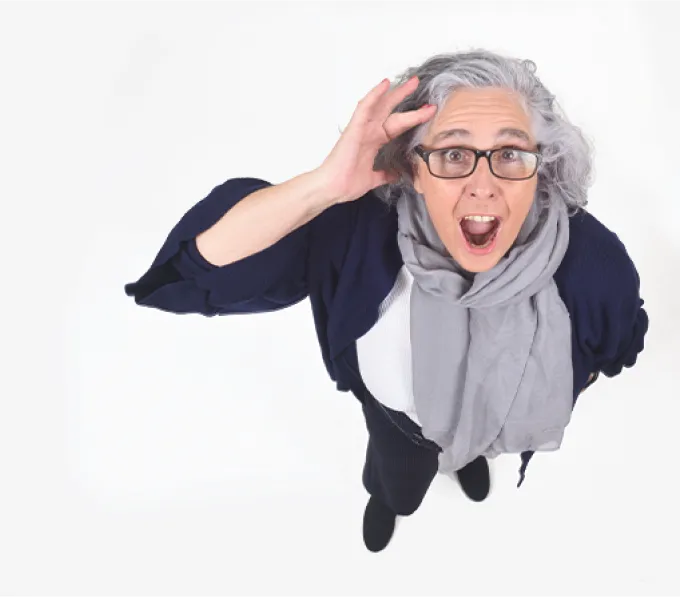 Surprised elderly woman with glasses looking up, holding her head in disbelief or excitement.