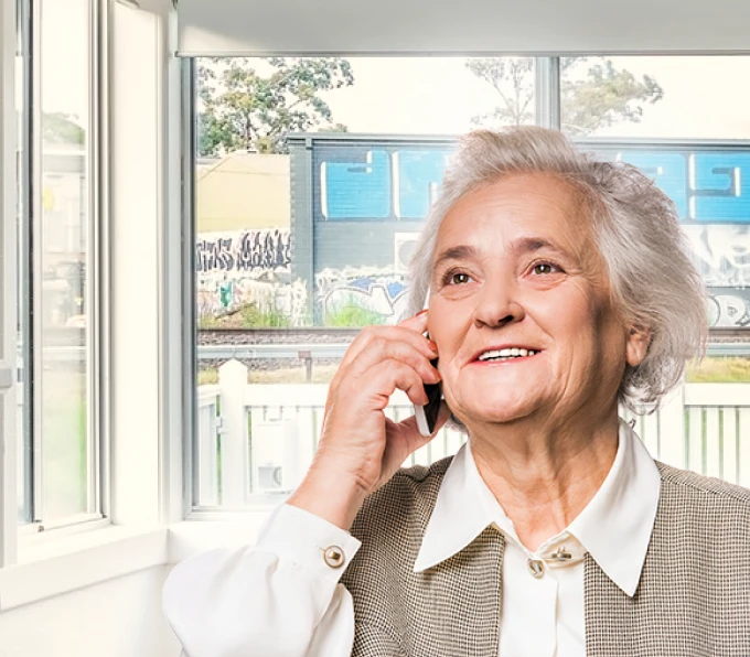 Elderly woman smiling and talking on the phone by a window overlooking a street with graffiti, appearing content and engaged.