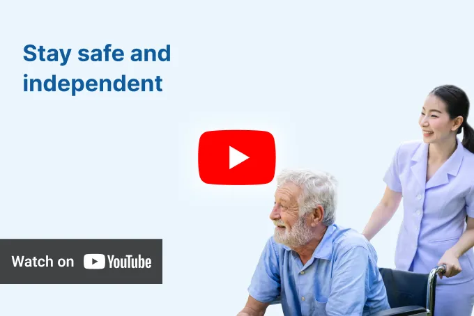 Elderly man in a wheelchair looking to the side with a smiling Asian caregiver standing behind him, beside text 'Stay safe and independent' and a 'Watch on YouTube' button.