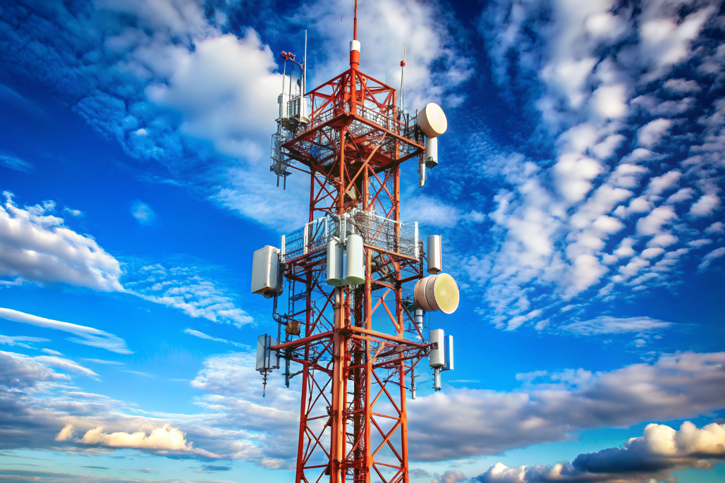 Red and white telecommunications tower with antennas against a vibrant blue sky and clouds.