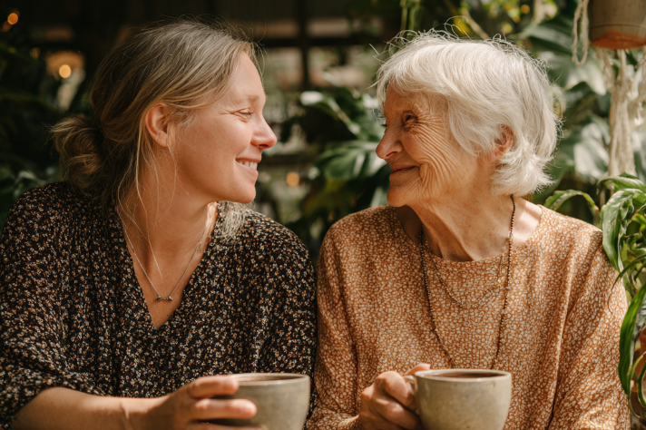 Adult daughter and elderly mother sharing a warm moment together over coffee, both smiling as they engage in conversatio