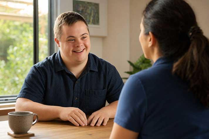 Young man with Down syndrome sitting at a dining table with a support worker across from him, both relaxed in conversation, warm afternoon light