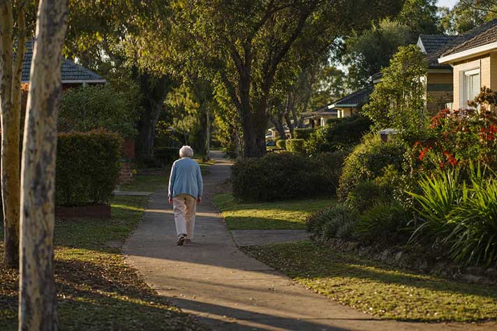 An elderly woman walking alone along a quiet suburban footpath, seen from behind at a distance, afternoon light